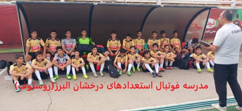 عکس گروهی از یک تیم فوتبال نوجوانان در کرج، شامل 14 بازیکن با لباس‌های زرد و مشکی با لوگوی "Rosto"، نشسته زیر سایه‌بان زمین ورزشی، در کنار یک مربی با لباس خاکستری و نشان "Coach"، در حال دریافت آموزش، با متن فارسی "مدرسه فوتبال استعدادهای درخشان" در پایین، نمایش‌دهنده محیط آموزشی و تیمی در فوتبال